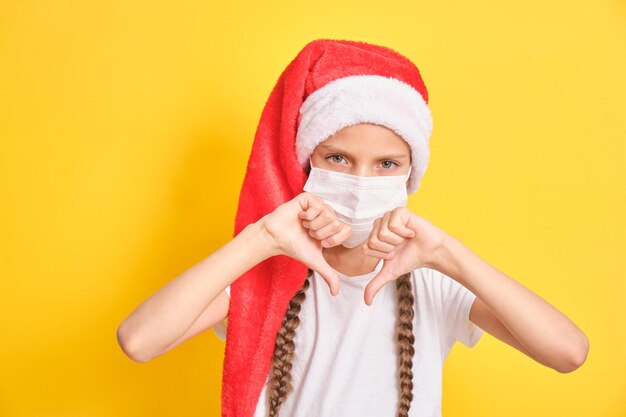 Adolescente con sombrero rojo de santa claus para la celebración navideña muestra el gesto de los pulgares hacia abajo sobre fondo amarillo, el niño está triste por el bloqueo durante la época navideña