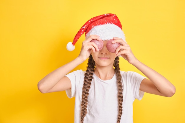 Adolescente con sombrero de santa claus sosteniendo bolas de navidad sobre fondo amarillo