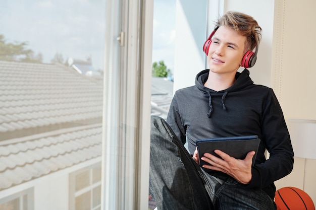 Adolescente sonriente con auriculares y tableta digital sentado en el alféizar de la ventana y mirando al cielo cuando se queda en casa durante el bloqueo