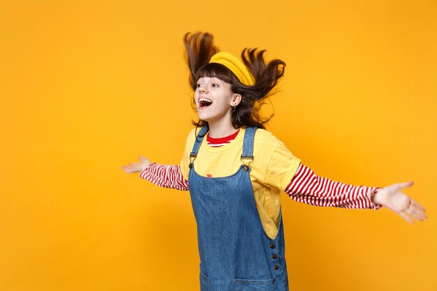 Una adolescente sonriente con boina francesa saltando con el pelo suelto mirando a un lado, extendiendo las manos aisladas en un fondo amarillo en el estudio. Emociones sinceras de la gente, concepto de estilo de vida. Simulacros de espacio de copia.