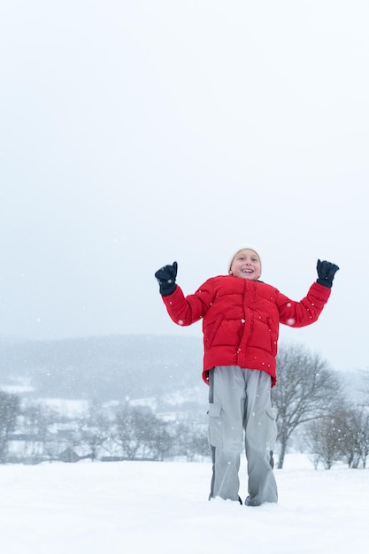 Un adolescente sonriente con una chaqueta roja salta durante la nevada Un niño con las manos en alto y disfruta de las vacaciones de invierno