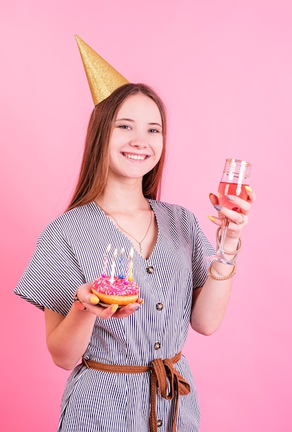 Adolescente sonriente cumpleañera con sombrero de cumpleaños sosteniendo dos donas con velas aislado en rosa