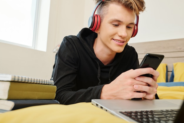 Adolescente sonriente escuchando música en auriculares y jugando en el teléfono inteligente cuando descansa en su habitación