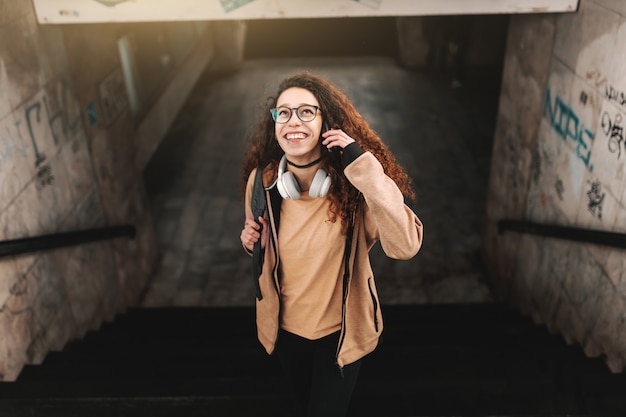 Adolescente sonriente con el pelo largo y rizado subiendo las escaleras en la estación de tren. Auriculares alrededor del cuello