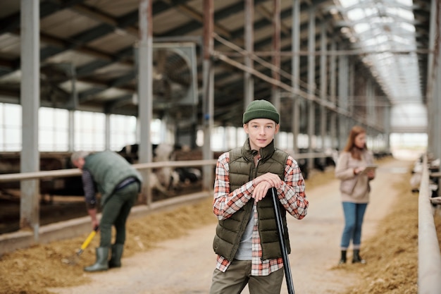 Adolescente sonriente en ropa de trabajo mirándote mientras está de pie contra el pasillo entre potreros con vacas lecheras y sus padres trabajando
