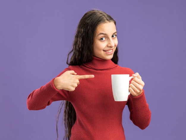 Foto adolescente sonriente sosteniendo y apuntando a una taza de té mirando a la cámara aislada en la pared púrpura