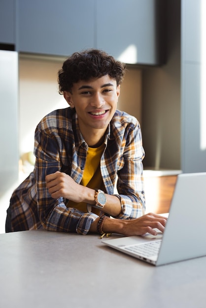 Un adolescente sonriente usando una computadora portátil en casa con una camisa a cuadros y un reloj