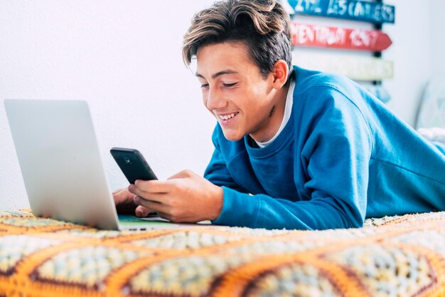 Foto un adolescente sonriente usando el teléfono móvil sobre la computadora portátil en la cama en casa