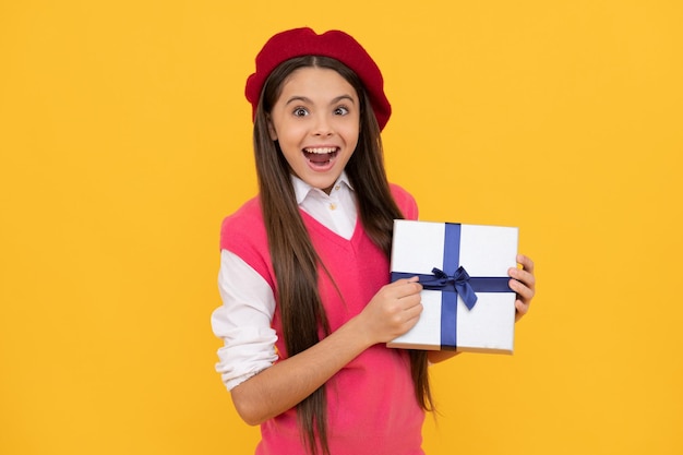 Una adolescente sorprendida con una boina francesa sostiene un regalo o una caja de regalo con fondo amarillo, de compras.