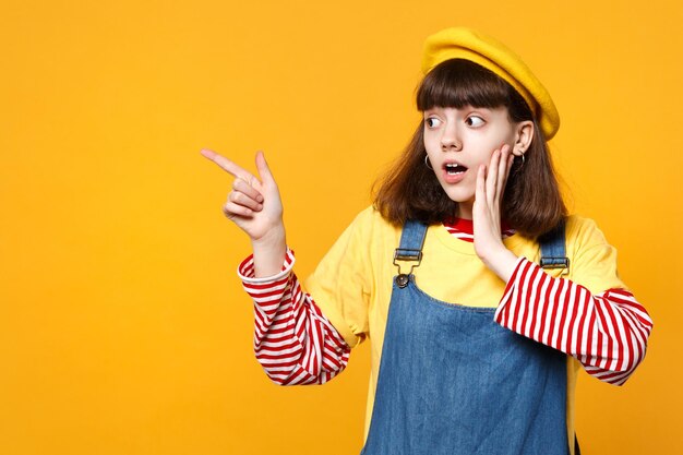 Adolescente sorprendida con boina francesa, vestido de mezclilla poniendo la mano en la mejilla, señalando con el dedo índice a un lado aislado en el fondo amarillo. Emociones sinceras de la gente, concepto de estilo de vida. Simulacros de espacio de copia.