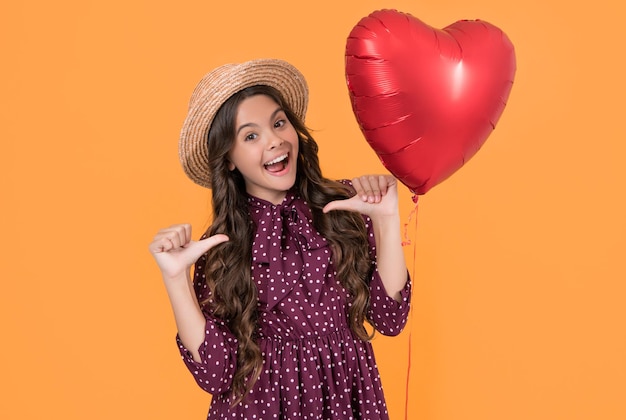 Una adolescente sorprendida con un globo de corazón rojo sobre un fondo amarillo se señala a sí misma