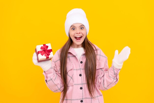 Una adolescente sorprendida con sombrero y mitones sostiene una caja de regalo sobre fondo amarillo año nuevo