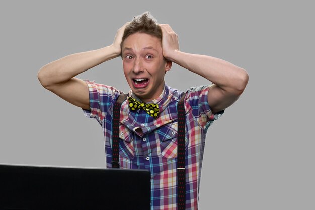 Adolescente sorprendido aturdido sobre fondo gris. Adolescente emocional cogidos de la mano en la cabeza mientras está sentado frente a la computadora portátil.