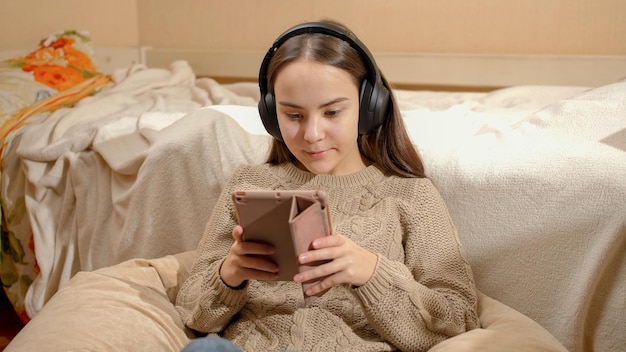 Adolescente sorridente ouvindo música em fones de ouvido usando computador tablet em seu quarto