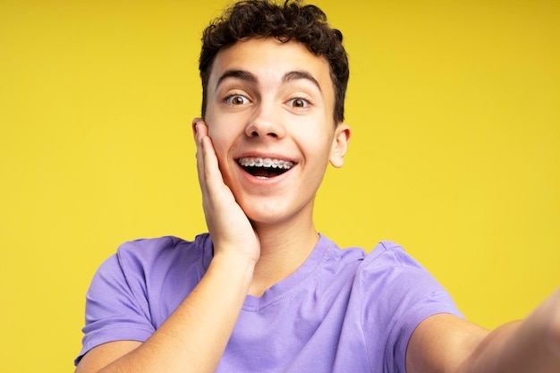 Adolescente sorridente usando aparelho dentário nos dentes tirando selfie isolado em fundo amarelo