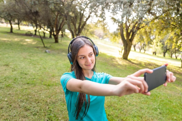 Adolescente sorridente vestindo uma camiseta turquesa e fones de ouvido, tirando uma selfie com seu celular