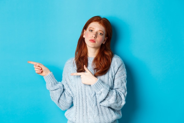 Adolescente temperamental com cabelo vermelho, apontando os dedos para a esquerda no logotipo, olhando incomodada e entediada, em pé sobre um fundo azul.