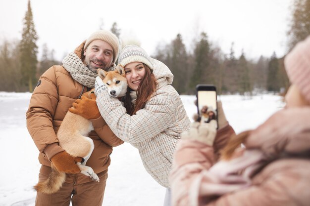 Adolescente tirando foto dos pais enquanto desfrutam de um passeio ao ar livre juntos na floresta de inverno, foco no casal adulto feliz segurando o cachorro