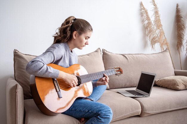 Una adolescente tocando la guitarra durante el aprendizaje electrónico frente a una computadora portátil en casa en el sofá