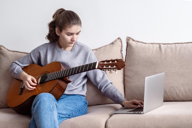 Una adolescente tocando la guitarra durante el aprendizaje electrónico frente a una computadora portátil en casa en el sofá