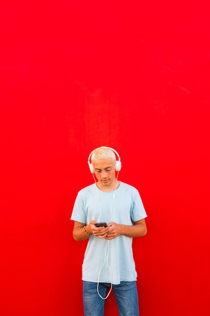 Adolescente u hombre usando su teléfono y escuchando música con auriculares con una pared de colores rojos en el fondo - concepto de estilo de vida musical - milenario y adulto en línea