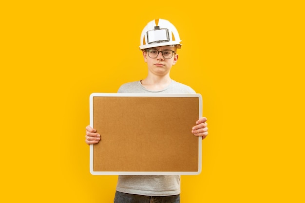 Adolescente usa anteojos y casco protector sosteniendo un tablero de corcho en blanco sobre fondo amarillo Copiar espacio Maqueta