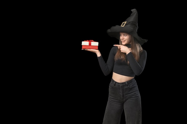 Una adolescente vestida de negro y un sombrero puntiagudo sostiene un regalo en la palma de la mano y lo señala con el dedo Aislado en el espacio de copia de fondo negro