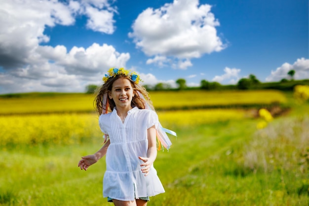 Adolescente con vestido blanco y corona ucraniana corre a través de campos amarillos y prados verdes contra el cielo nublado