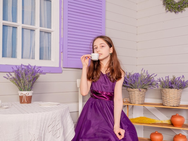 Una adolescente con un vestido morado bebe té en una taza blanca. La niña está sentada a la mesa.