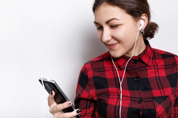 Adolescente vestindo camisa xadrez elegante, ouvindo sua música favorita com fones de ouvido sorrindo, olhando para a tela. Estudante bonita em fones de ouvido em pé contra branco