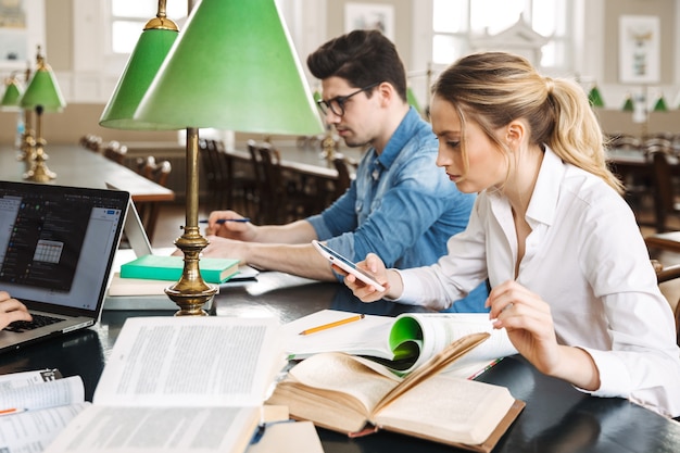 Adolescentes alegres inteligentes que estudian juntos en la biblioteca, leyendo libros, usando computadoras portátiles