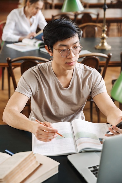 Adolescentes alegres inteligentes que estudian juntos en la biblioteca, leyendo libros, usando computadoras portátiles