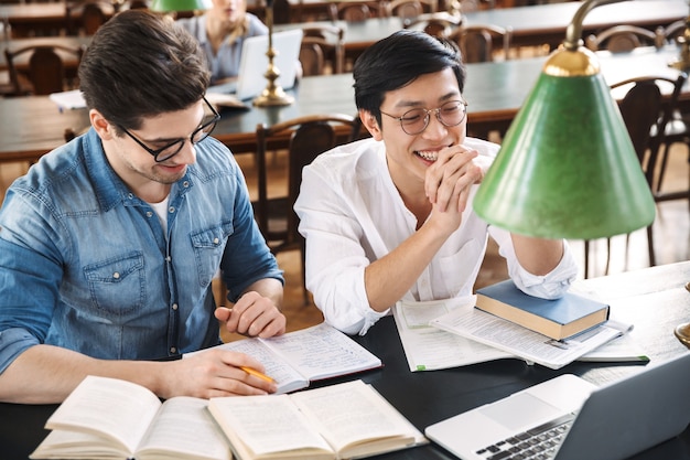 Adolescentes alegres inteligentes que estudian juntos en la biblioteca, leyendo libros, usando computadoras portátiles