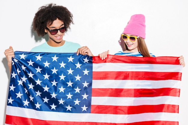 Adolescentes americanos. Casal jovem e descolado segurando a bandeira americana na frente deles e sorrindo em pé contra um fundo branco