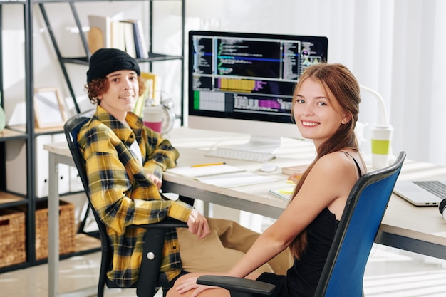 Adolescentes sonrientes inteligentes sentados en el escritorio en casa, haciendo los deberes y mirando a la cámara