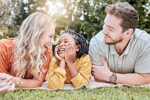 Adopción familiar y picnic en una manta en el parque con la madre, el padre y la niña negra en la naturaleza, relájese y feliz. Los niños y los padres de la diversidad se relajan en el césped en el patio y disfrutan del verano con amor o una sonrisa.