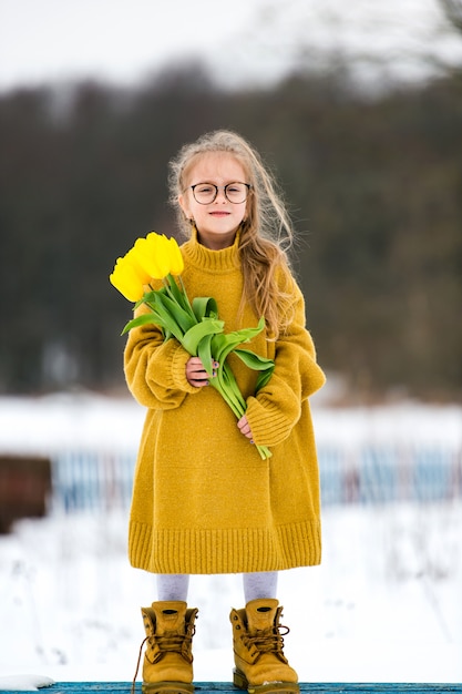 Adorabe kleines Mädchen, das erwachsene übergroße Strickjacke und Stiefel des großen Vaters trägt. Schönes junges Mädchen in den Gläsern, die auf Bank mit Blumenstrauß von gelben Tulpen am Tag des verschneiten Winters stehen.