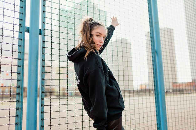 Una adorable y adorable chica con el pelo oscuro y un jersey negro posa en la plaza del deporte en un cálido día de primavera