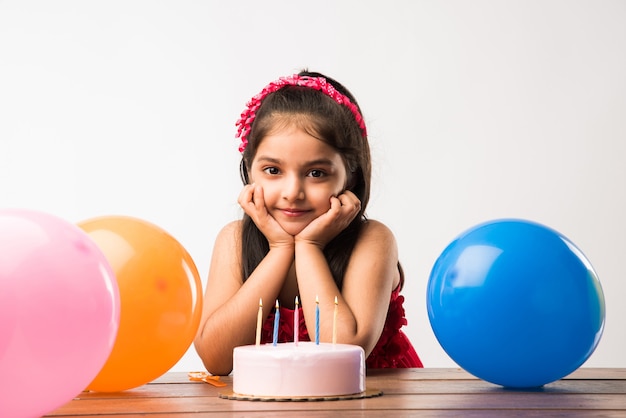 Adorable adorable niña india o asiática celebrando un cumpleaños mientras sostiene el pastel de fresa y sopla velas en la mesa o de pie aislado sobre fondo blanco o rojo