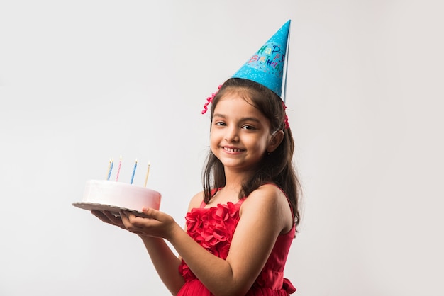 Adorable adorable niña india o asiática celebrando un cumpleaños mientras sostiene el pastel de fresa y sopla velas en la mesa o de pie aislado sobre fondo blanco o rojo