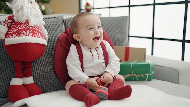 Adorable bebé hispano sonriendo confiado sentado en el sofá junto al árbol de Navidad en casa