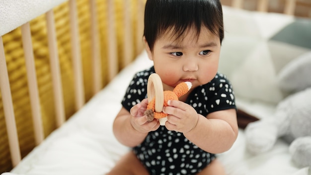 Adorable bebé hispano sosteniendo juguete sentado en la cuna en el dormitorio