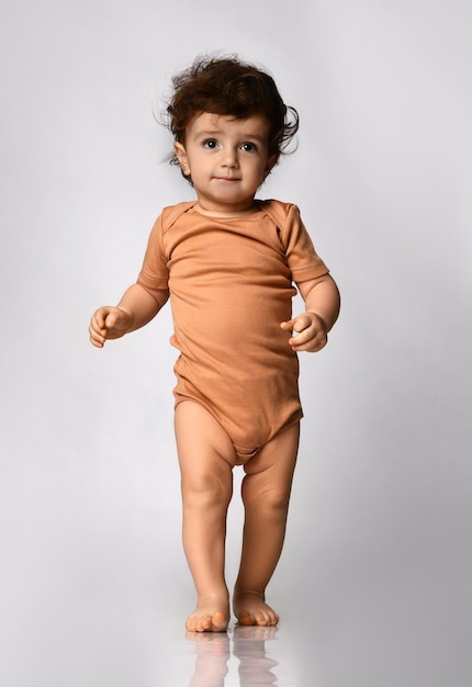 Adorable bebé rizado con traje beige da sus primeros pasos en un fondo gris