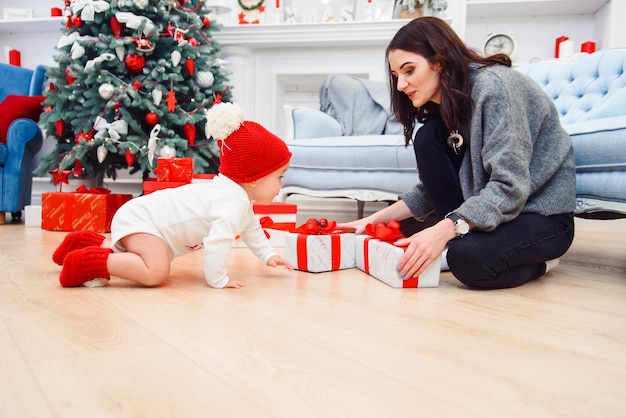 Adorable bebé sonriente arrastrándose sobre el parquet a los regalos de Navidad cerca de su madre, que se sienta en el suelo.