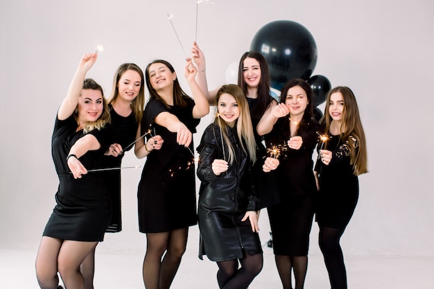 Adorable caucásica siete mujeres posando con luces de bengala y globos de aire. Hermosas chicas en vestidos negros con bengalas y riendo en la fiesta de cumpleaños.