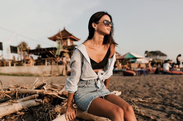 Adorable chica encantadora con pelo corto oscuro en falda y camisa azul sentada en la playa de arena en la luz del sol