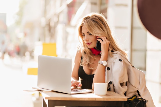 Adorable chica rubia con pañuelo rojo escribiendo en el teclado mientras habla por teléfono con un amigo. Mujer joven elegante en reloj de pulsera con ordenador portátil en la cafetería al aire libre después de una taza de té.