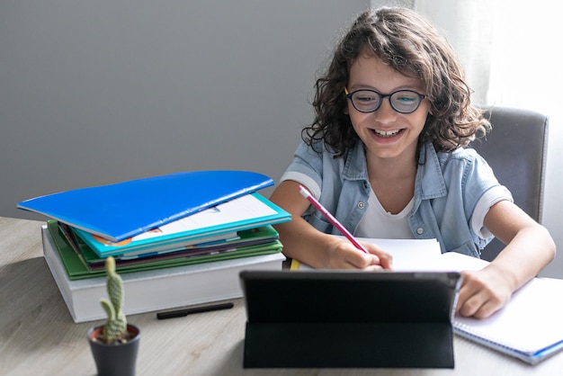 Un adorable colegial con anteojos estudiando en línea desde casa usando una tableta digital