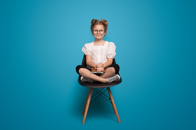 Adorable colegiala está sonriendo a la cámara a través de gafas sentado en un sillón sosteniendo algunos libros en ...