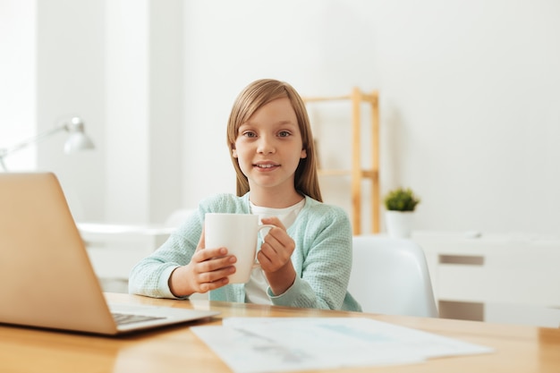 Adorable y encantadora niña comprometida a ponerse cálida y acogedora mientras leía un artículo y trabajaba en su tarea en casa